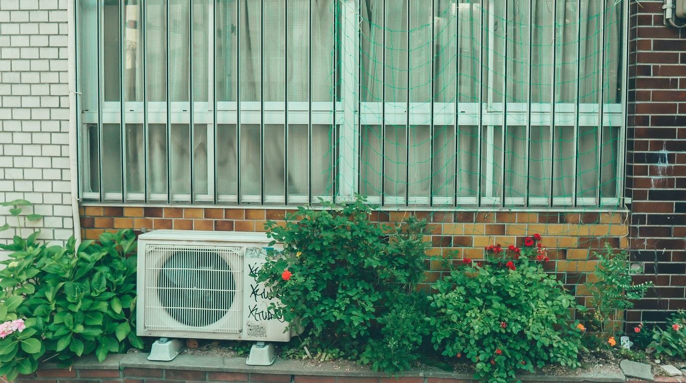 에어컨 이전설치 - 벽돌 건물 창문 아래 화단 옆에 놓인 에어컨 실외기